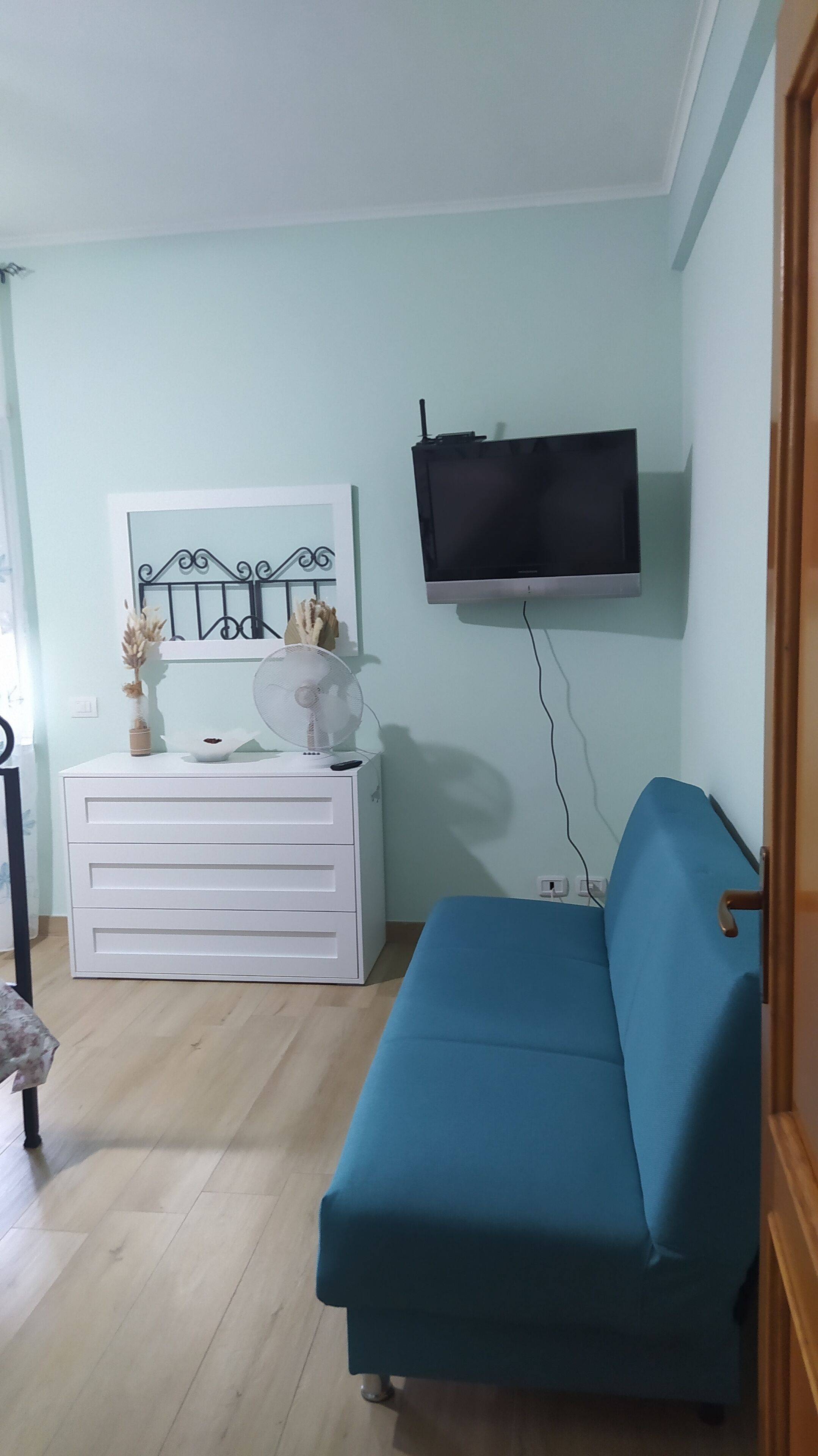 Un salon avec un canapé bleu, une commode blanche, un téléviseur à écran plat fixé au mur et un ventilateur posé sur la commode.