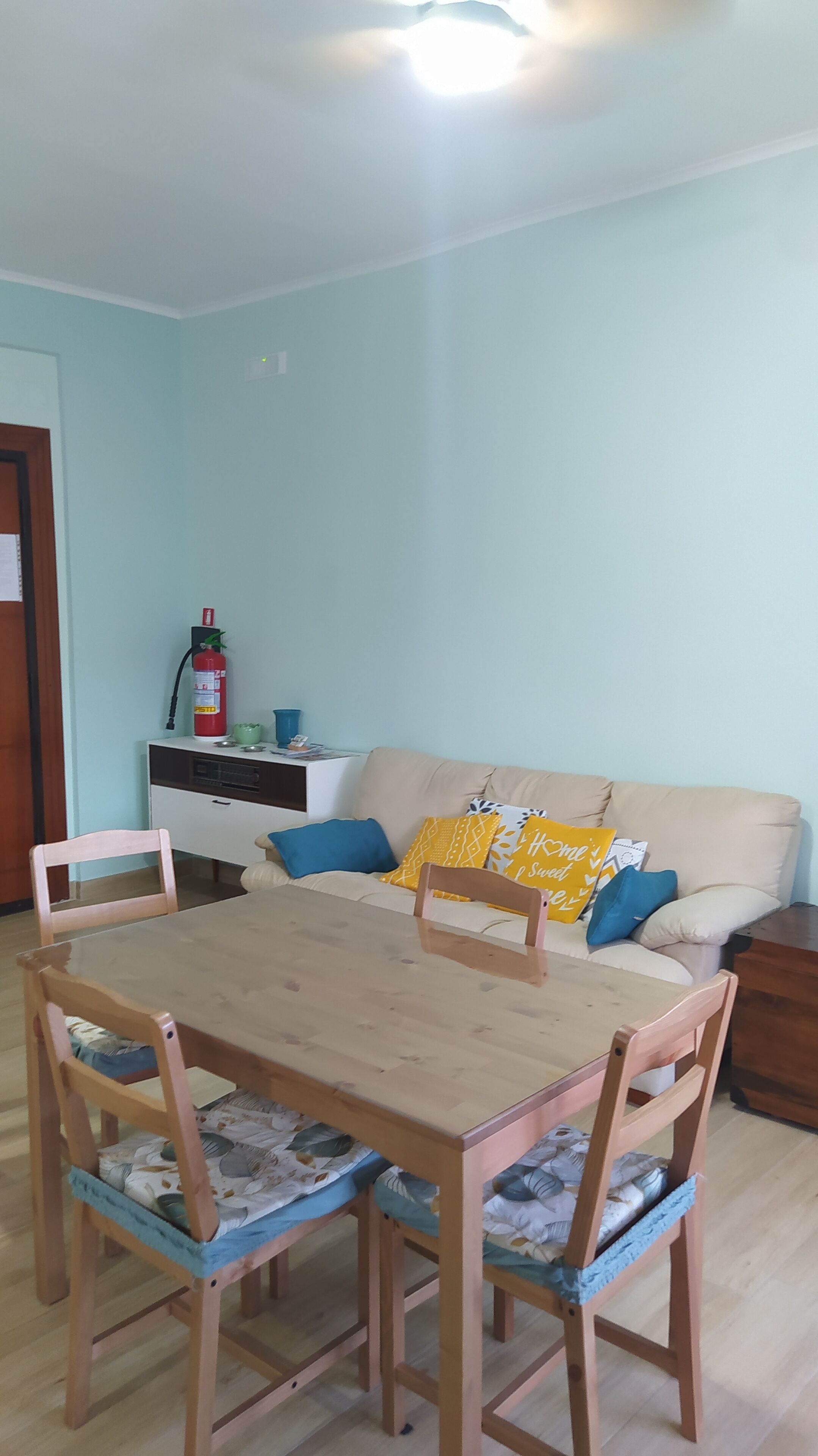 Un coin repas avec une table et des chaises en bois, un canapé agrémenté de coussins à motifs et un extincteur placé sur un meuble.