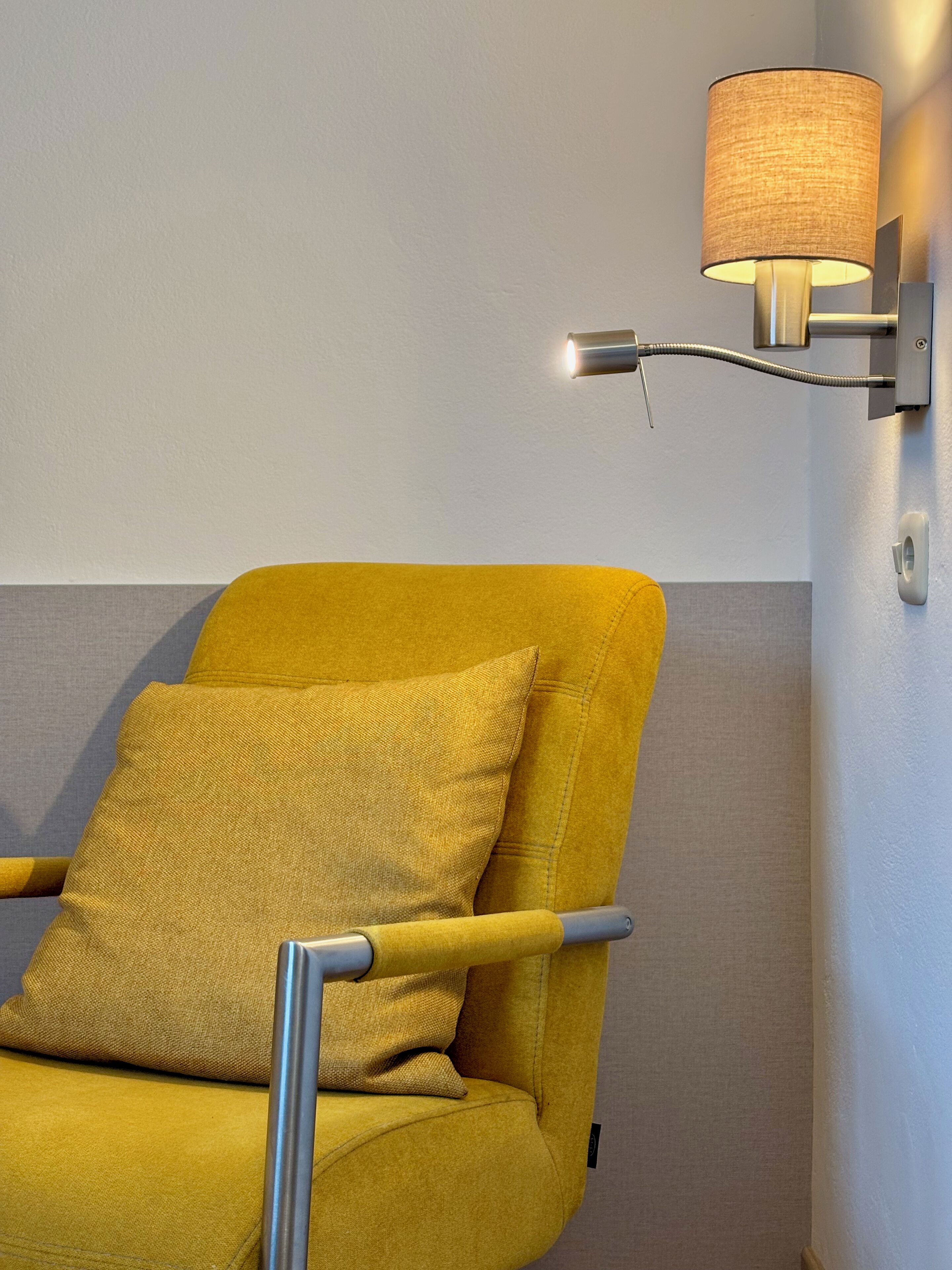 A yellow armchair with a cushion, a wall-mounted lamp, and a grey wall.