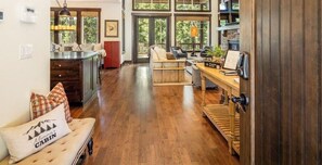 A spacious living area with a wooden floor, a sofa with a cushion, a dining table, and a kitchen area with a window.