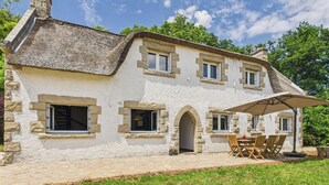 Un bâtiment en pierre avec un toit de chaume, un patio couvert doté de sièges extérieurs et un ciel d’un bleu éclatant.