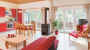 Ein gemütliches Wohnzimmer mit einer Couch, einem Esstisch und Stühlen. Es gibt eine Küchenzeile mit roten Schränken und einem Herd. Ein Fernseher steht auf einem Holztisch, und eine Pflanze ist in einem roten Topf.