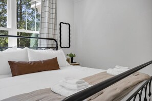 Un dormitorio con cama, almohadas blancas, una almohada marrón, cabecera de madera, ventana con vista a árboles y una obra de arte abstracto negra en la pared.