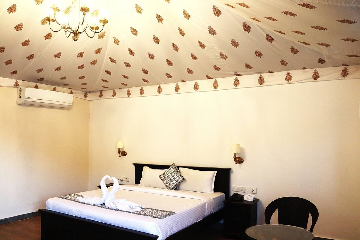 A hotel room with a bed, a chandelier, an air conditioning unit, a nightstand, a chair, and a table.
