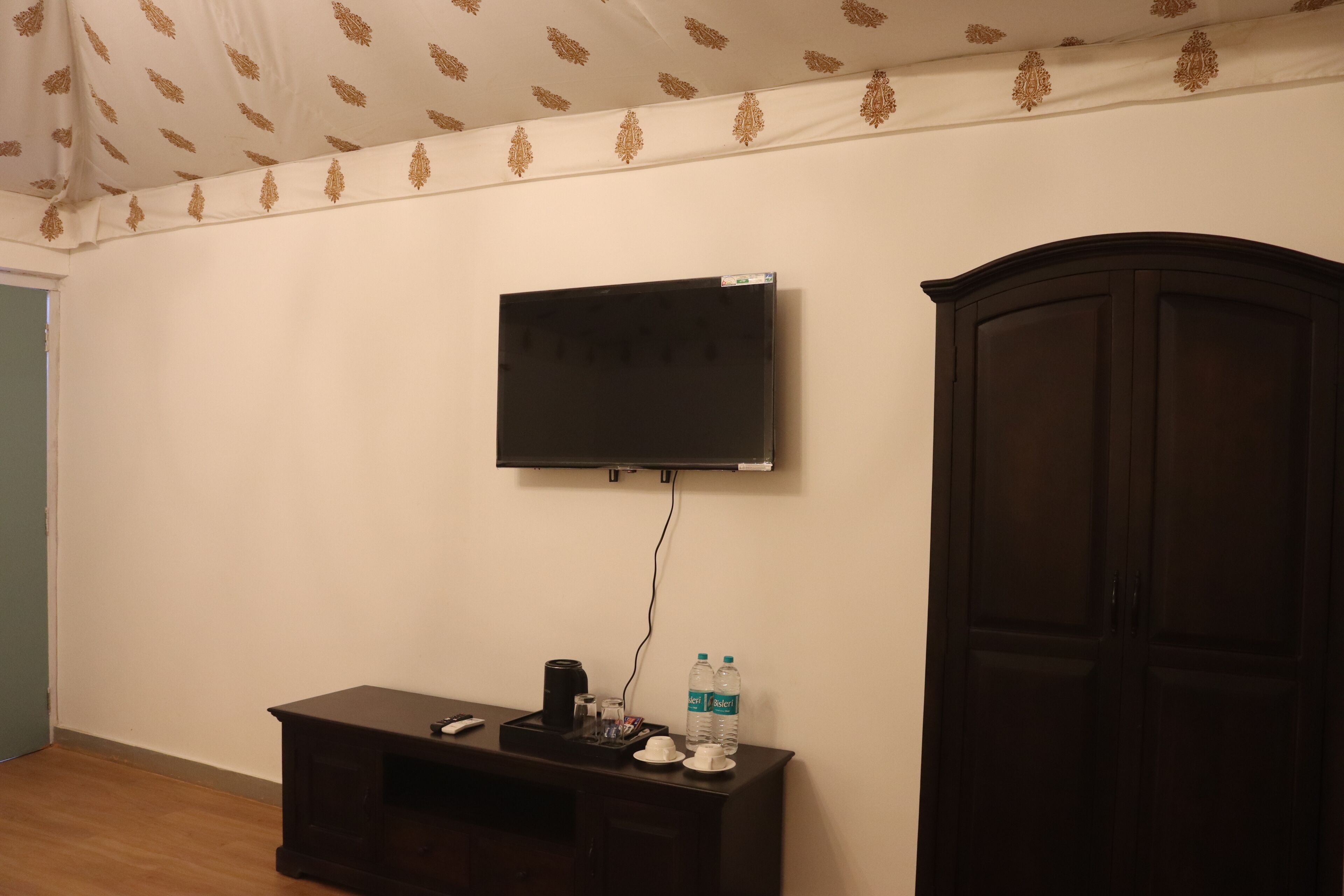 A room with a flat-screen TV mounted on the wall, a small wooden desk with a remote, a water bottle, and a cabinet.