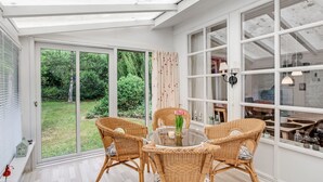 Ein heller Raum mit Glastür zu einem Garten, Korbmöbel und einem runden Tisch mit einer Vase.