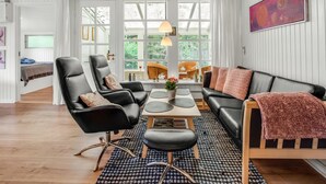 Ein modernes Wohnzimmer mit einem schwarzen Ledersofa, einem schwarzen Ledersessel, einem Holztisch und einem gemusterten Teppich. Ein Fenster mit weißen Vorhängen, ein Bild an der Wand und ein Deckenventilator sind ebenfalls zu sehen.