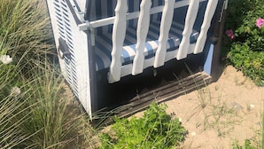 A beach chair with a striped pattern and a white seat, placed on sandy ground with grass and flowers around.