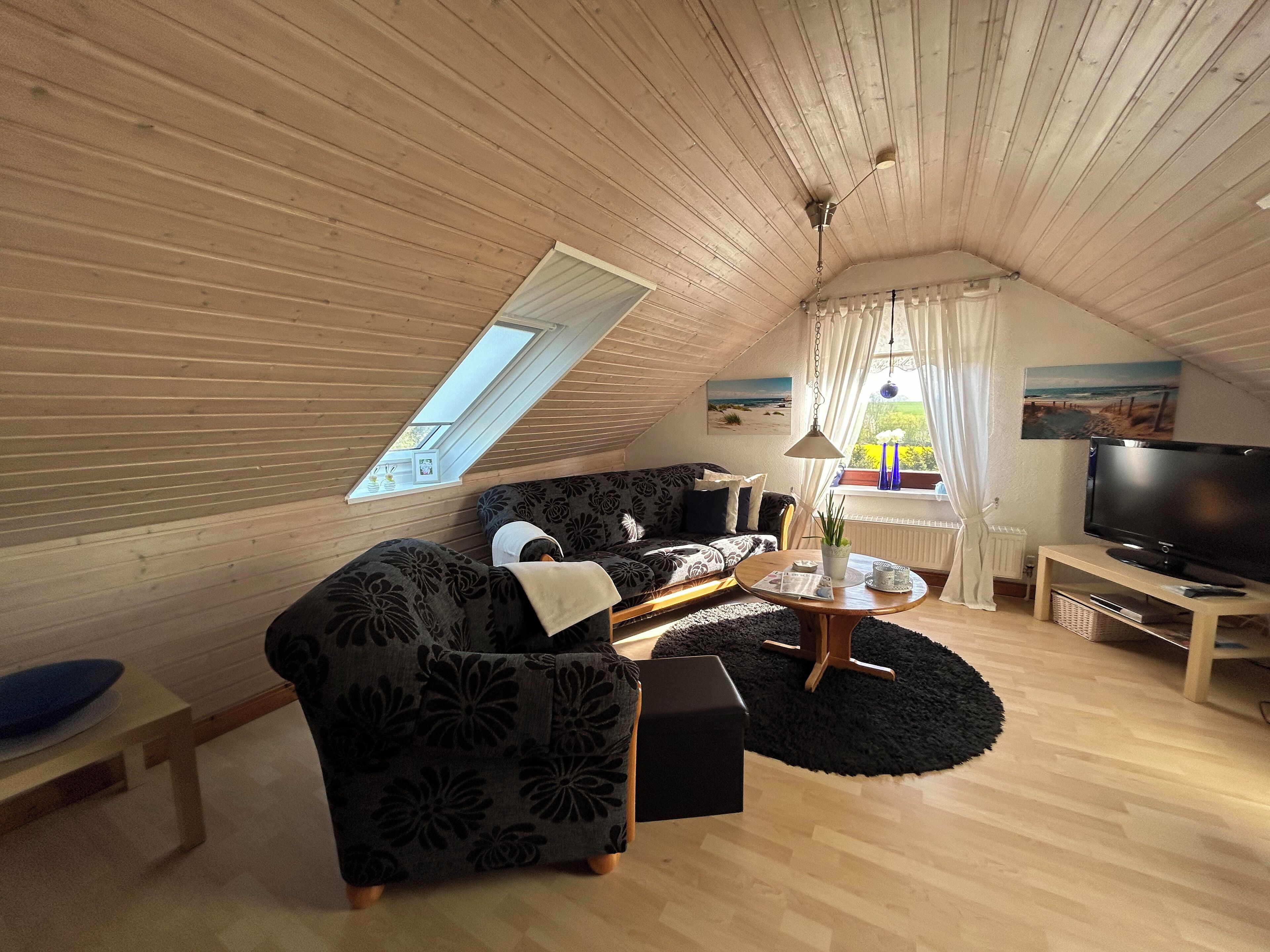 En hyggelig loftstue med højt til loftet, stort vindue, sofa, sofabord og et fladskærms-tv.