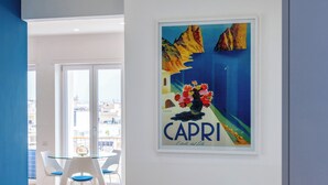 A modern dining area with a glass table and white chairs, a framed artwork of Capri, and a view of the city through large windows. - At Twenty-One: new apartment with exclusive panoramic views! (Napoli)