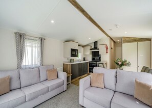A modern living room with two grey sofas, a kitchen with a black stove, and a wooden ceiling beam.
