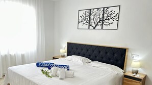 Una cama bien tendida con sábanas blancas, un almohadón azul con texto y una obra de arte con motivos de árbol en la pared.