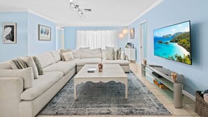 Una sala de estar con un sofá seccional color beige, una mesa de centro, un televisor de pantalla plana y un ventanal con vista a la playa.