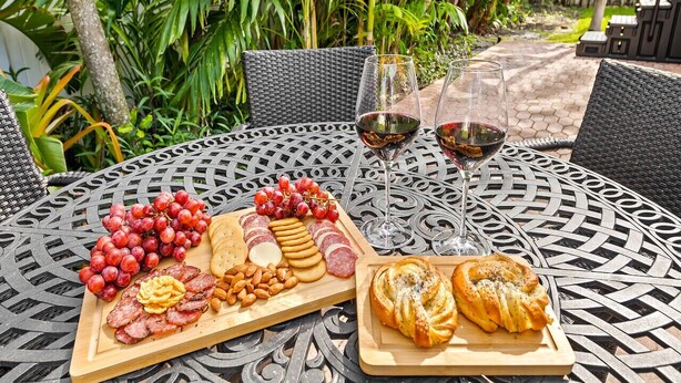 Una mesa redonda al aire libre con diseño de celosía metálica, decorada con una tabla de embutidos, dos copas de vino tinto y una silla de mimbre.