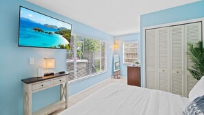Un dormitorio con cama, televisor montado en la pared, escritorio con lámpara y ventana con persianas.