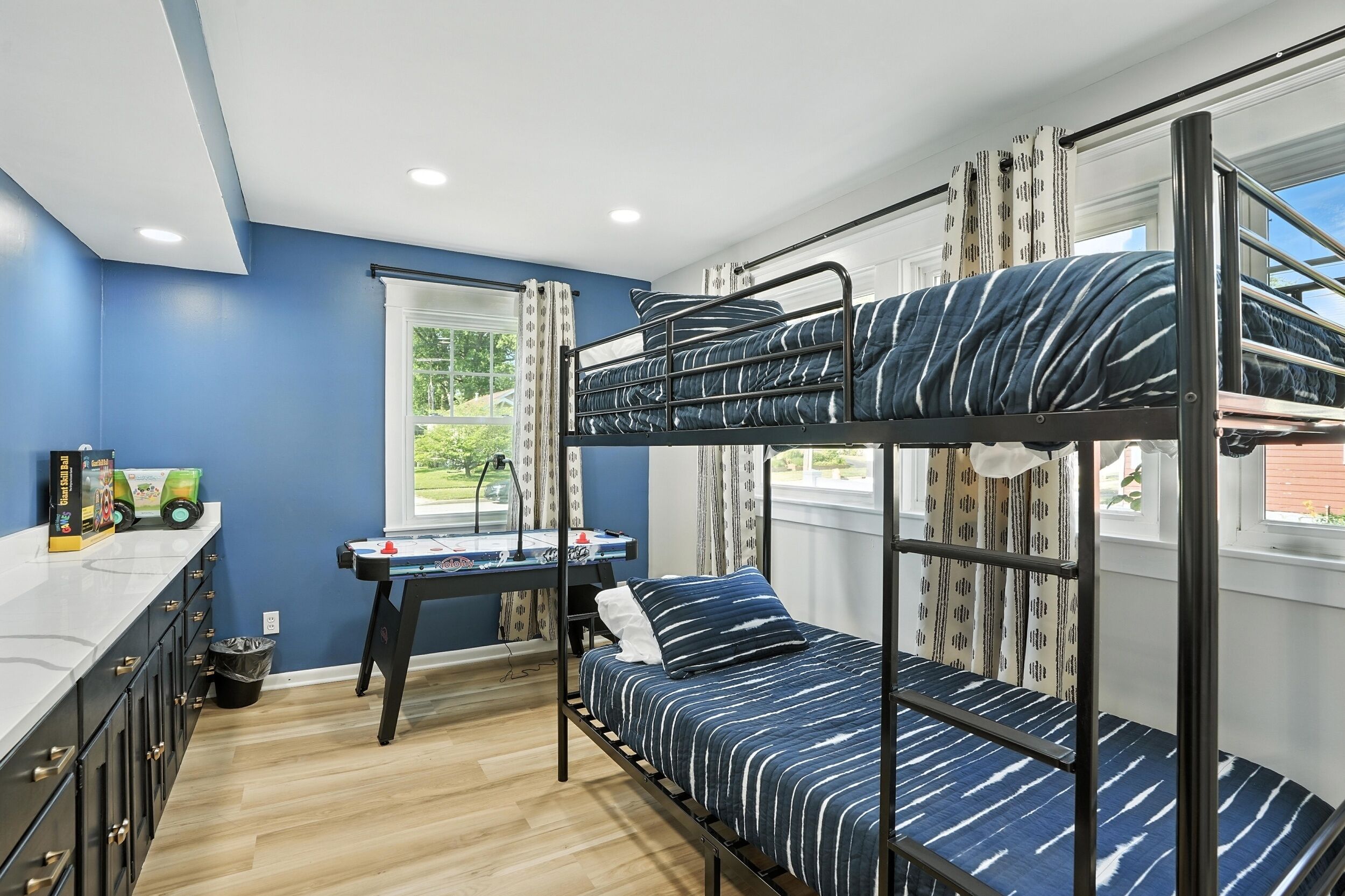 A bunk bed room with a desk, a window with curtains, and a view of greenery outside.