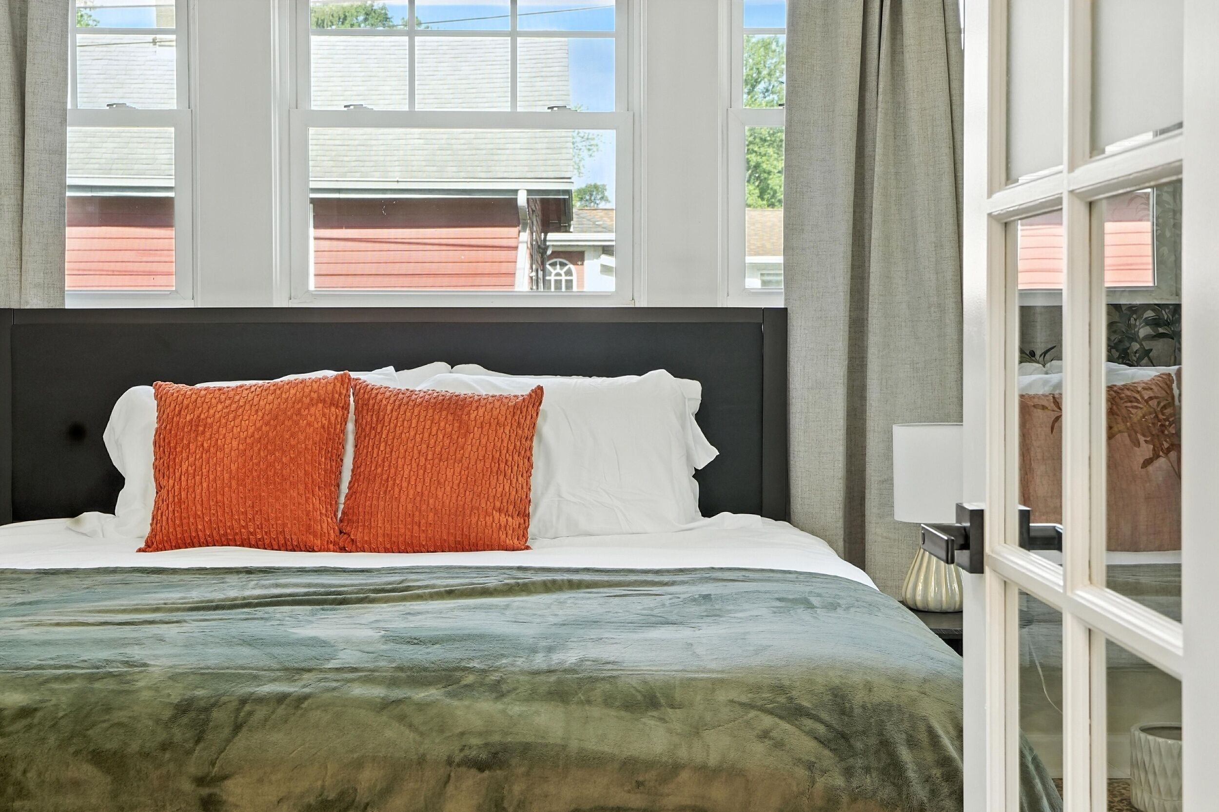 A neatly made bed with white and orange pillows, a green and blue blanket, and a view of buildings through large windows.