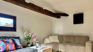 Ein Wohnzimmer mit grauem Sofa, bunten Kissen, einem weißen Beistelltisch, einem beigen Sofa, einem an der Wand montierten Fernseher und einem Fenster mit Blick auf ein Gebäude.