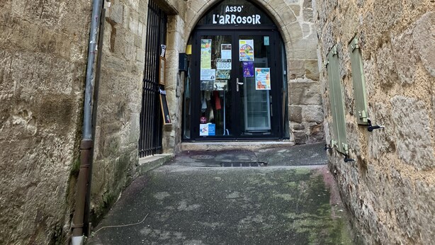 A narrow alleyway with stone walls, a black door labeled "Asso L'ArrosoiR", and a small window above the door.
