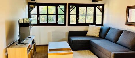 A living room with a sectional sofa, a small white table, a TV on a stand, and a window with multiple panes.