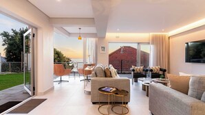 A modern living room with a sofa, coffee table, and chairs. There is a large window with a view of a building and a garden.