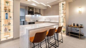 A modern kitchen with a marble island, bar stools, and glass shelving with decorative items.