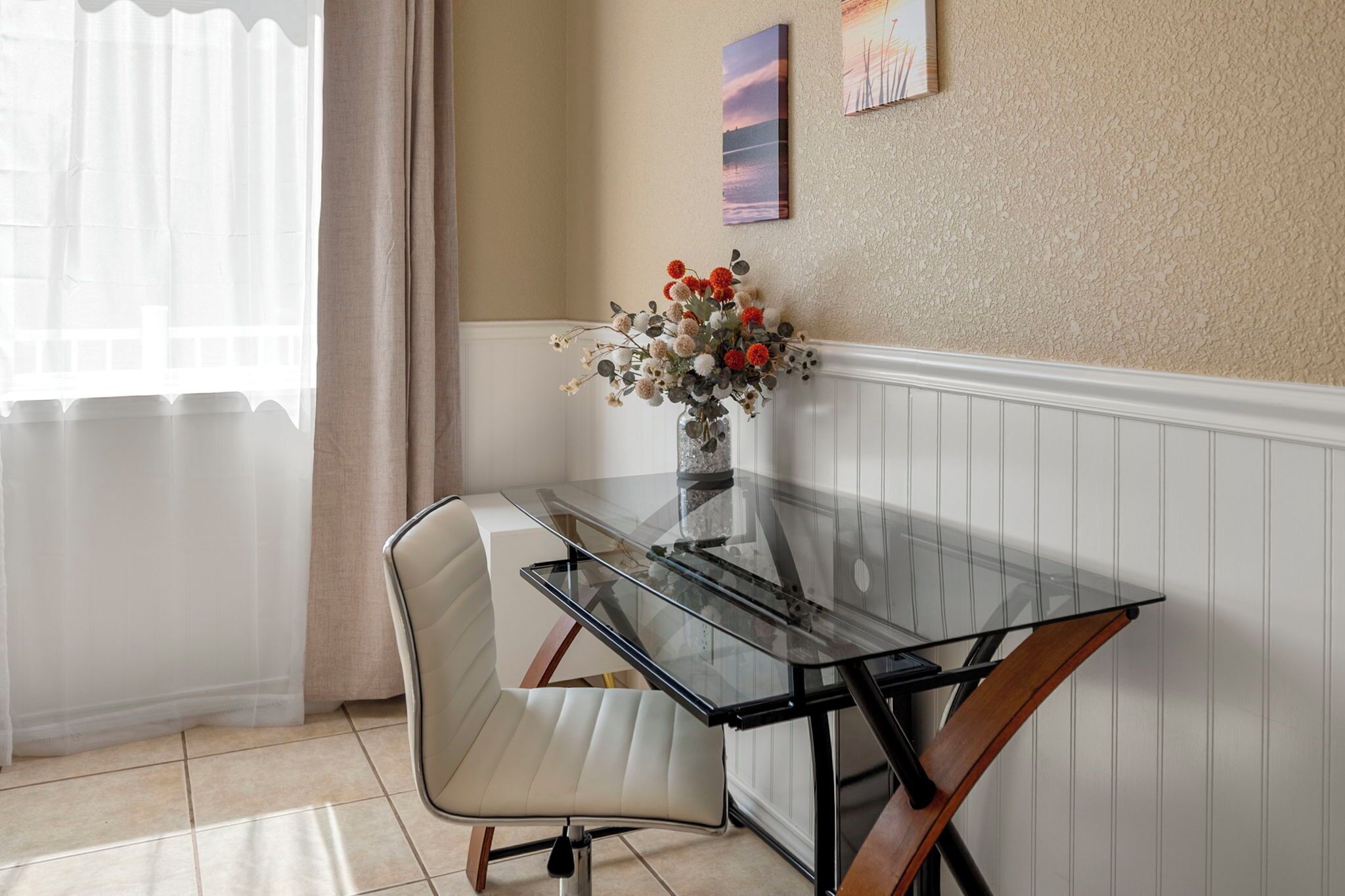 A dining area with a glass table, white cushioned chairs, a vase with flowers, and a wall with a painting.