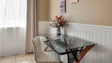 A dining area with a glass table, white cushioned chairs, a vase with flowers, and a wall with a painting.