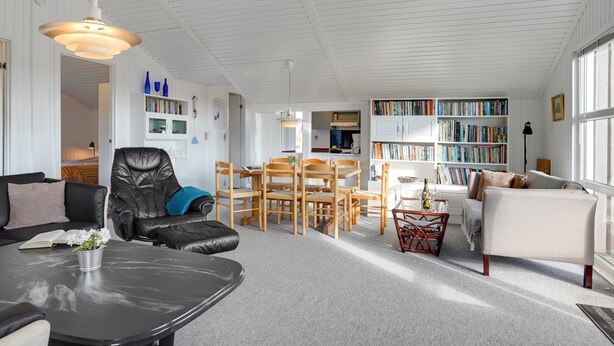 Een ruime woonkamer met een zwarte leren bank, een witte fauteuil, een eethoek met houten stoelen en een boekenplank vol boeken.
