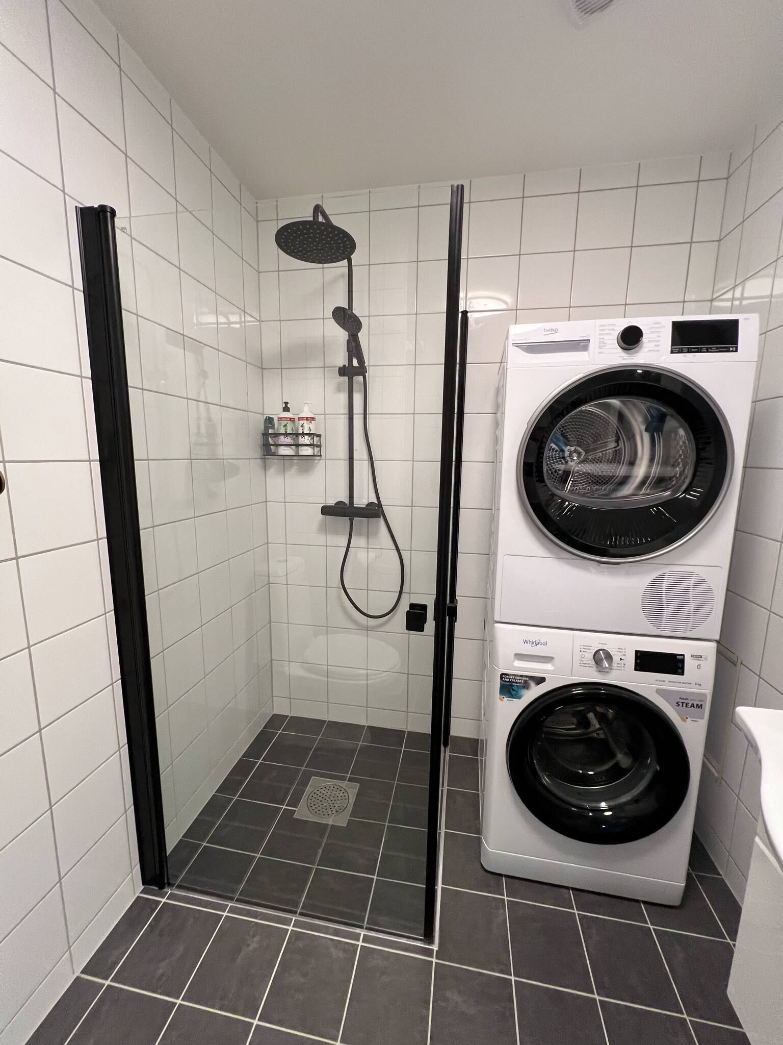A compact bathroom with a walk-in shower, a stacked washer and dryer, and a small shelf with toiletries.
