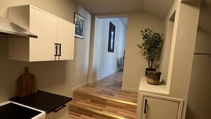 Una cocina compacta con estufa, horno y armarios blancos. Una escalera de madera conduce a un pasillo con una puerta y una planta en maceta.