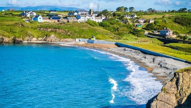 A coastal scene with a clear blue sea, a rocky shoreline, and a small village with houses and buildings in the background. - The Granary Loft at Annestown House (Annestown)