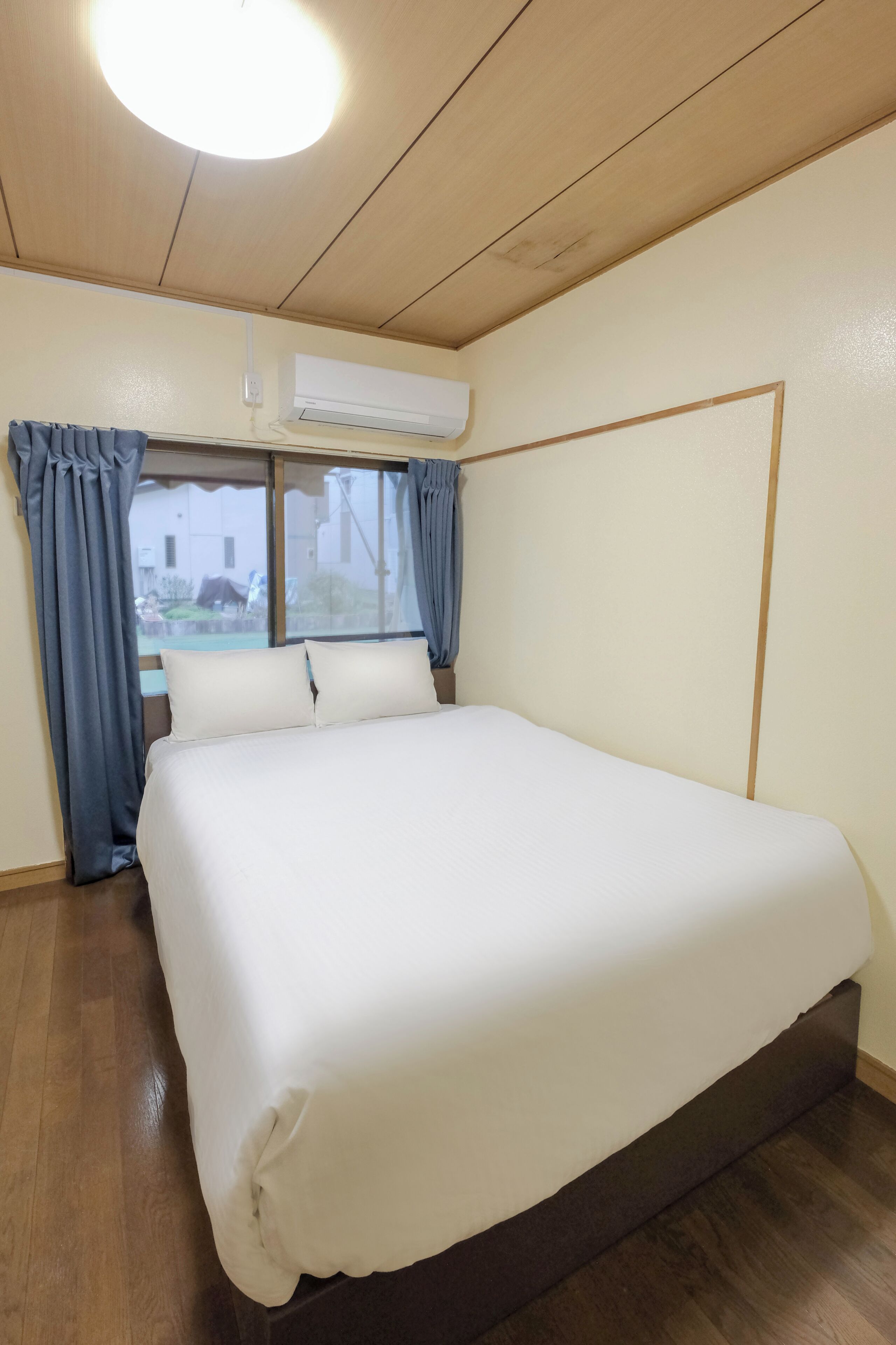 Una habitación de hotel con una cama grande, suelo de madera, unidad de aire acondicionado y ventana con cortinas.