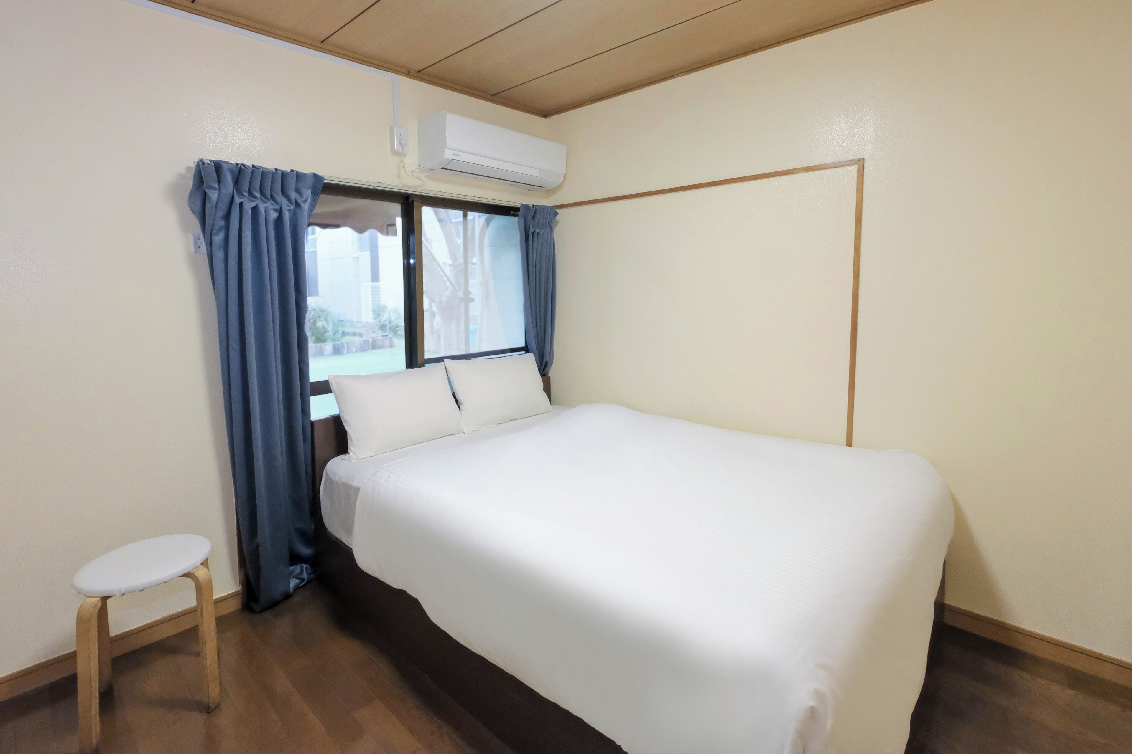 A room with a bed, a small stool, a window with curtains, and an air conditioning unit.