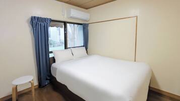 Una habitación con una cama, un taburete pequeño, una ventana con cortinas y un equipo de aire acondicionado.