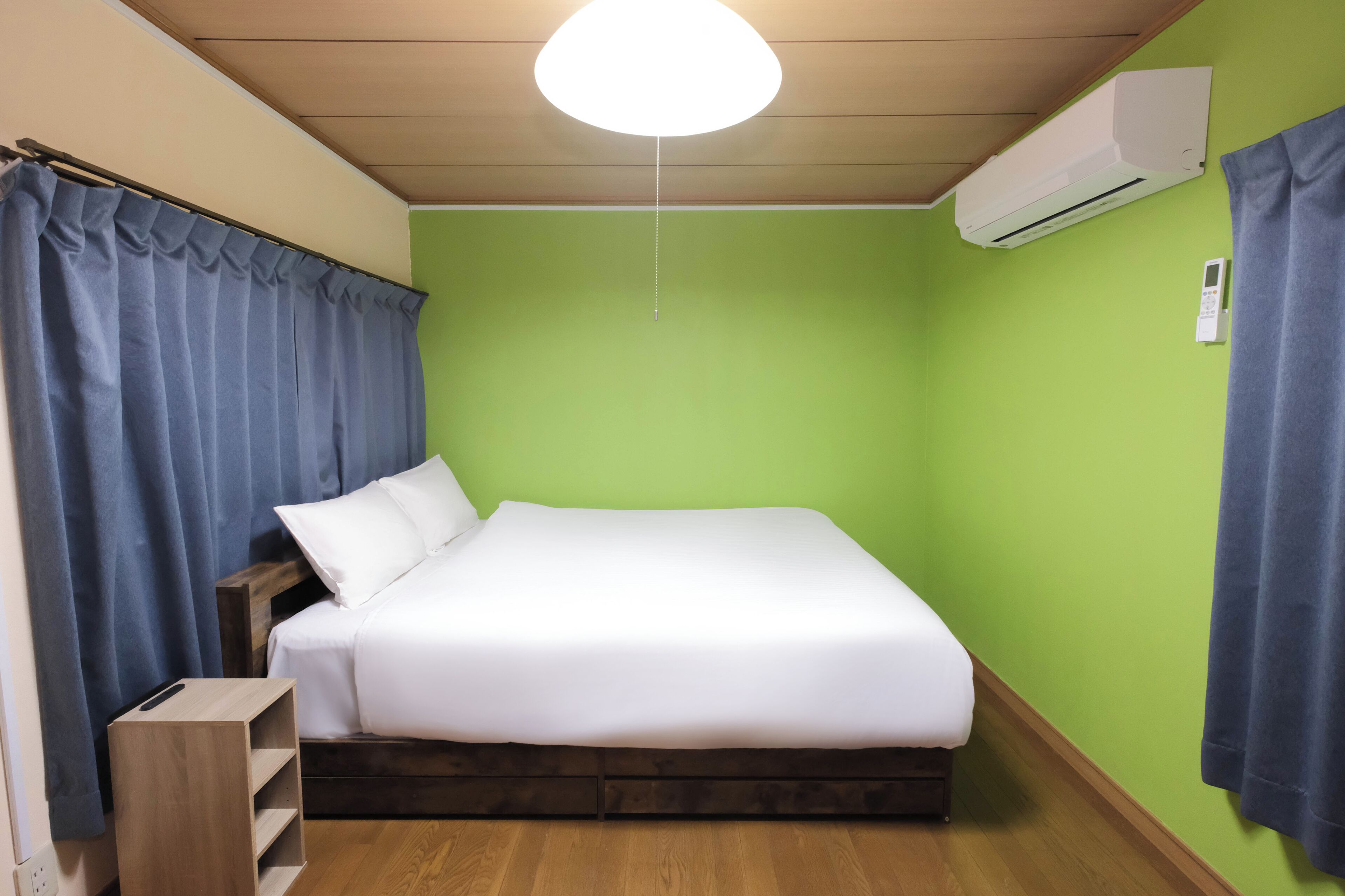 A single bed with white bedding, a wooden headboard, a nightstand, a blue curtain, and a wall-mounted air conditioner.
