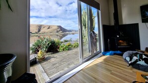 A room with a large sliding glass door leading to a deck with a view of a lake and mountains. - 3-bed gem in Birdlings Flat with stunning lake & ocean views – a rare retreat! (Little River)
