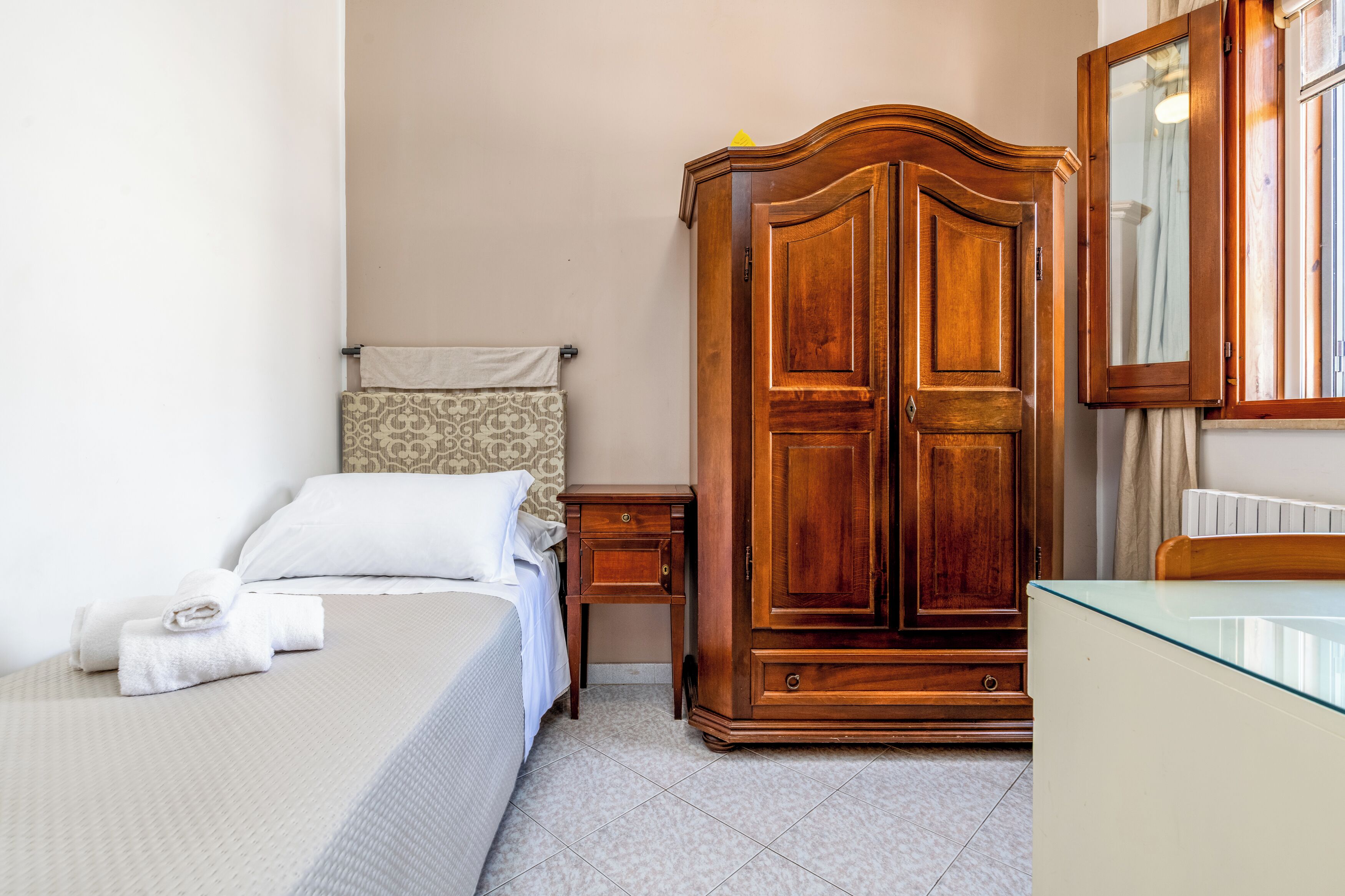 A hotel room with a bed, a wooden wardrobe, a bedside table, and a mirror.
