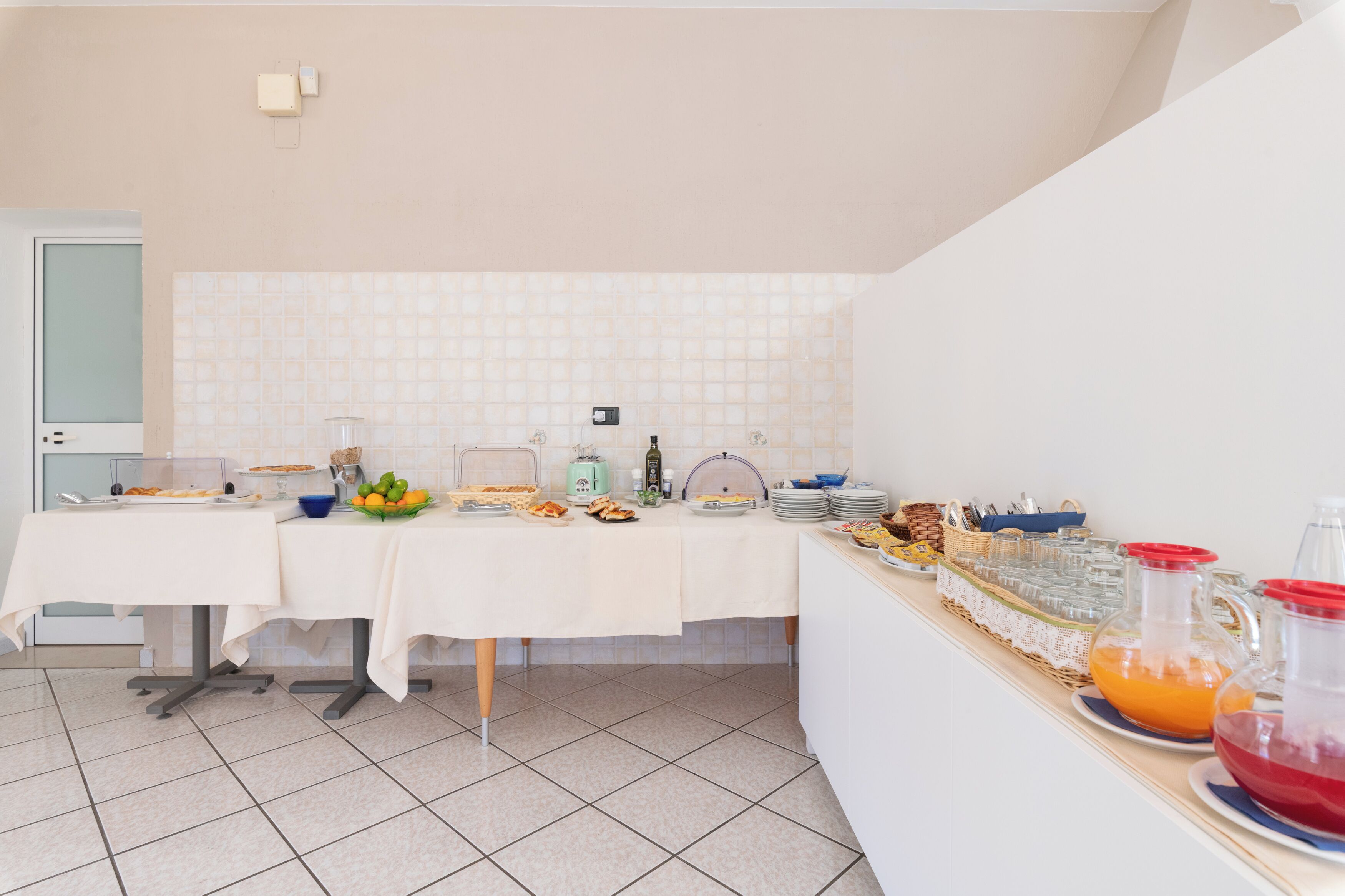 A breakfast buffet setup with various food items and beverages on a table, a tiled backsplash, and a white wall.