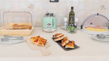 Una encimera de cocina con un tostador, una cesta de pan, una tabla de cortar con porciones de pizza, un plato con rebanadas de queso y un bol de jamón en rodajas.