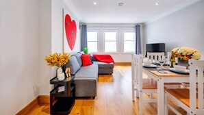 A modern living room with a grey sofa, a dining table set for two, and a large window with curtains.