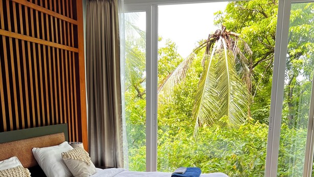 A bedroom with a large bed, wooden paneling on the wall, and a view of lush greenery through large windows. - Apartment with garden view (Tambon Karon)