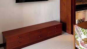 A modern living room with a flat-screen TV mounted on the wall, a wooden cabinet with drawers, and a patterned tablecloth on a dining table. - Apartment with garden view (Tambon Karon)