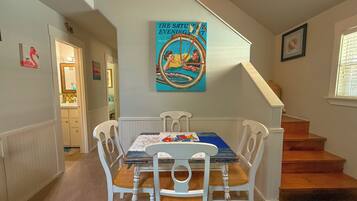 Un coin repas avec une table en bois et des chaises blanches, un tableau au mur et un escalier menant à l’étage.