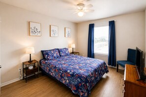 Ein Schlafzimmer mit einem Bett, zwei Nachttischen, einem Deckenventilator, einem Fenster mit Vorhängen und einem Fernseher.
