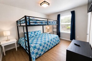 Een stapelbed met een gebedrukte spreideken, een nachtkastje met een lamp, een bureau met een afstandsbediening en een raam met gordijnen.