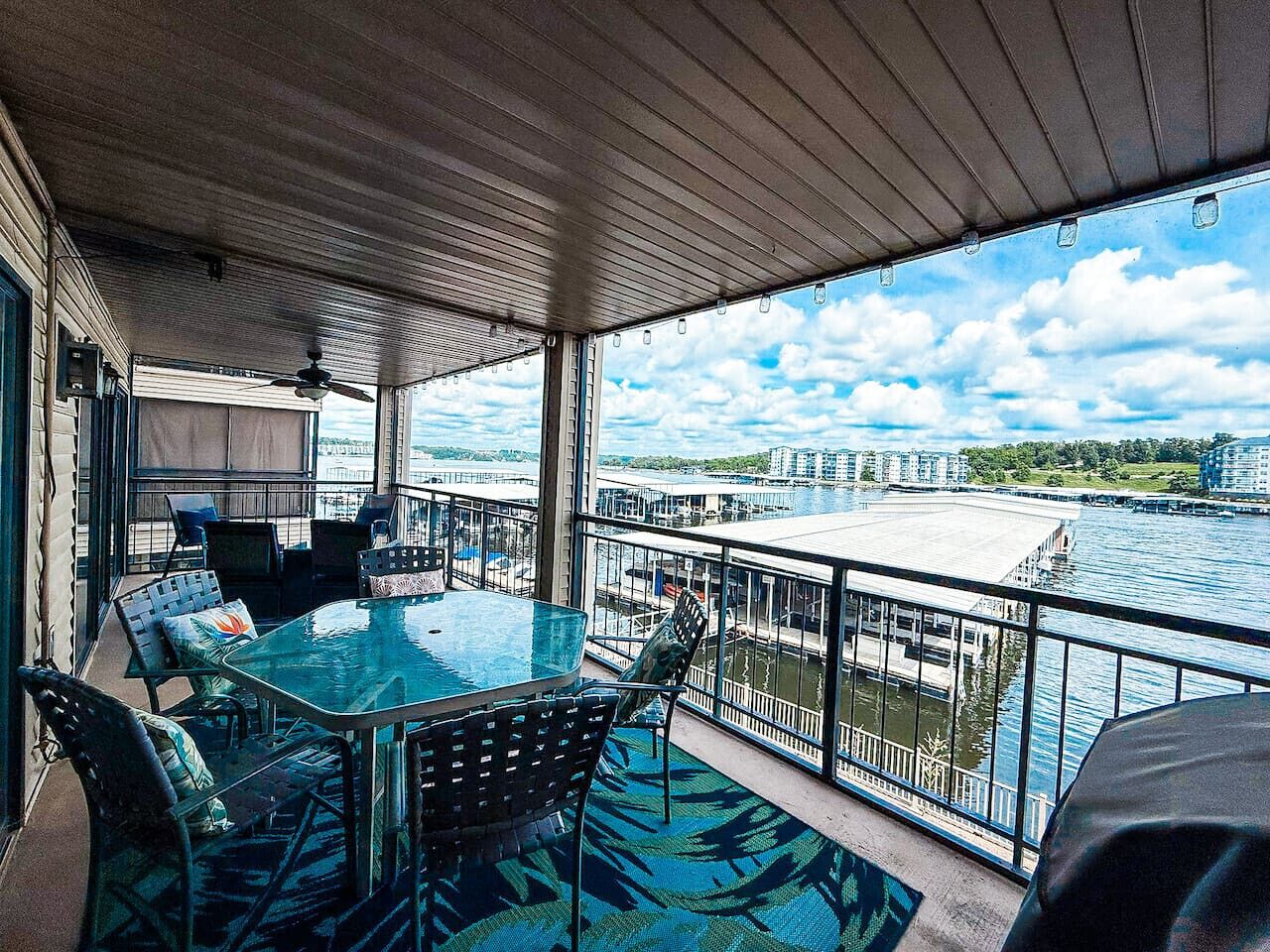 A covered outdoor balcony with a glass table and chairs, overlooking a body of water with buildings in the background.