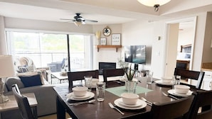 A dining area with a dark wooden table set for four, surrounded by dark chairs. There is a television mounted on the wall above the table. - Lakefront Luxury 2BD/2BA Condo at The Falls-7PP (Lake Ozark)