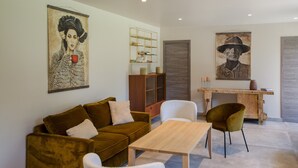 Una sala de estar con un sofá marrón, un sillón blanco y una mesa de centro de madera. Se observa una estantería y un cuadro en la pared.