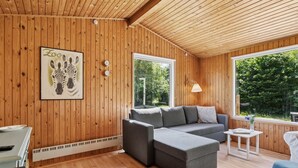 Interior acogedor de una cabaña con paredes de madera, un sofá gris, una mesita y un ventanal con vistas a la vegetación.